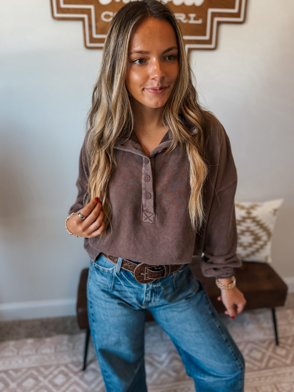 A woman wearing a brown long-sleeve henley shirt and blue jeans, standing in a home interior setting.