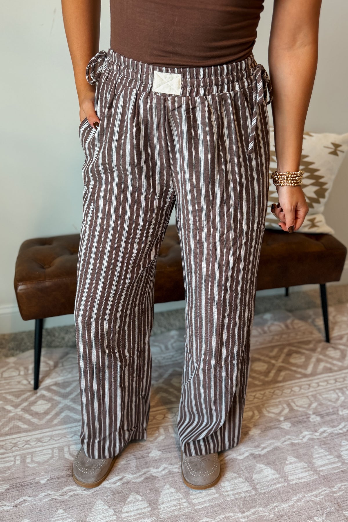 A person wearing brown and beige vertically striped wide-leg pants, paired with a brown top and brown shoes, standing in a room with a patterned rug and a brown bench.