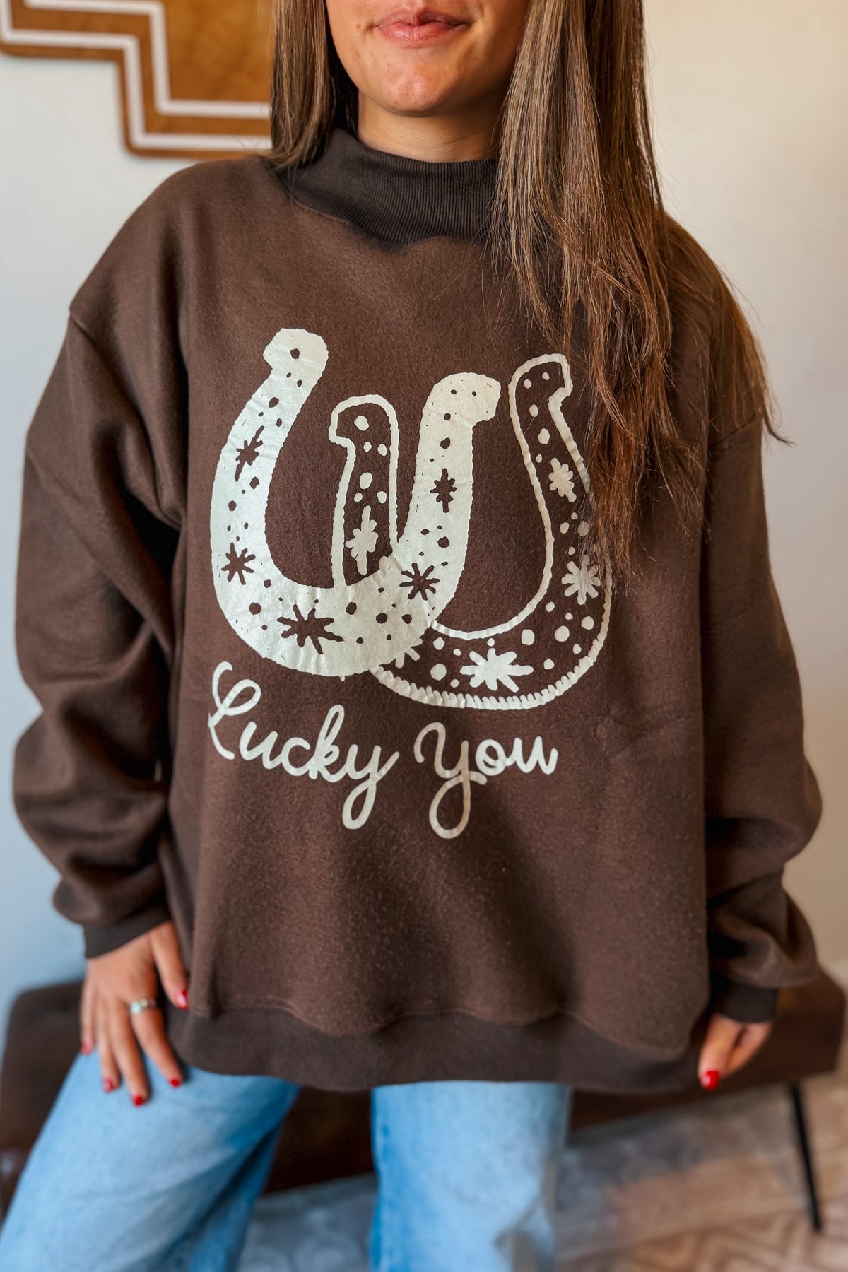 A woman wearing a brown crewneck sweatshirt with a white graphic of a horseshoe decorated with stars and the phrase 'Lucky You' printed below it, paired with light blue jeans.