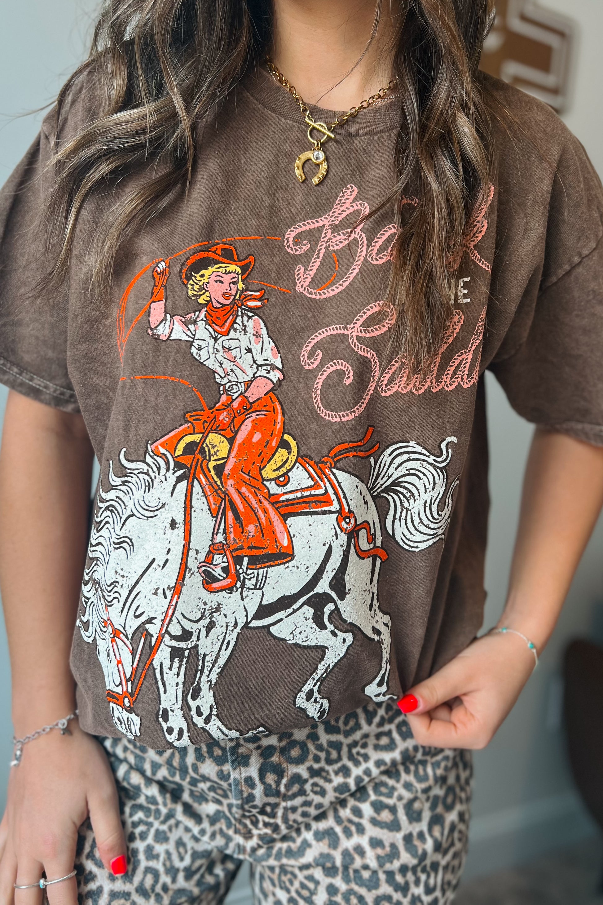 Brown vintage-wash graphic tee featuring a cowgirl riding a horse with “Back in the Saddle” text, styled with leopard print bottoms.