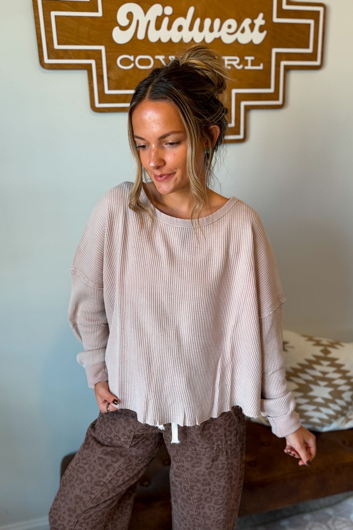 A woman wearing a light beige ribbed knit long-sleeve top and brown leopard-print pants, standing indoors sign.