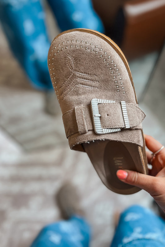Taupe suede western mule with silver buckle strap, studded toe detail, and low-profile slip-on design.