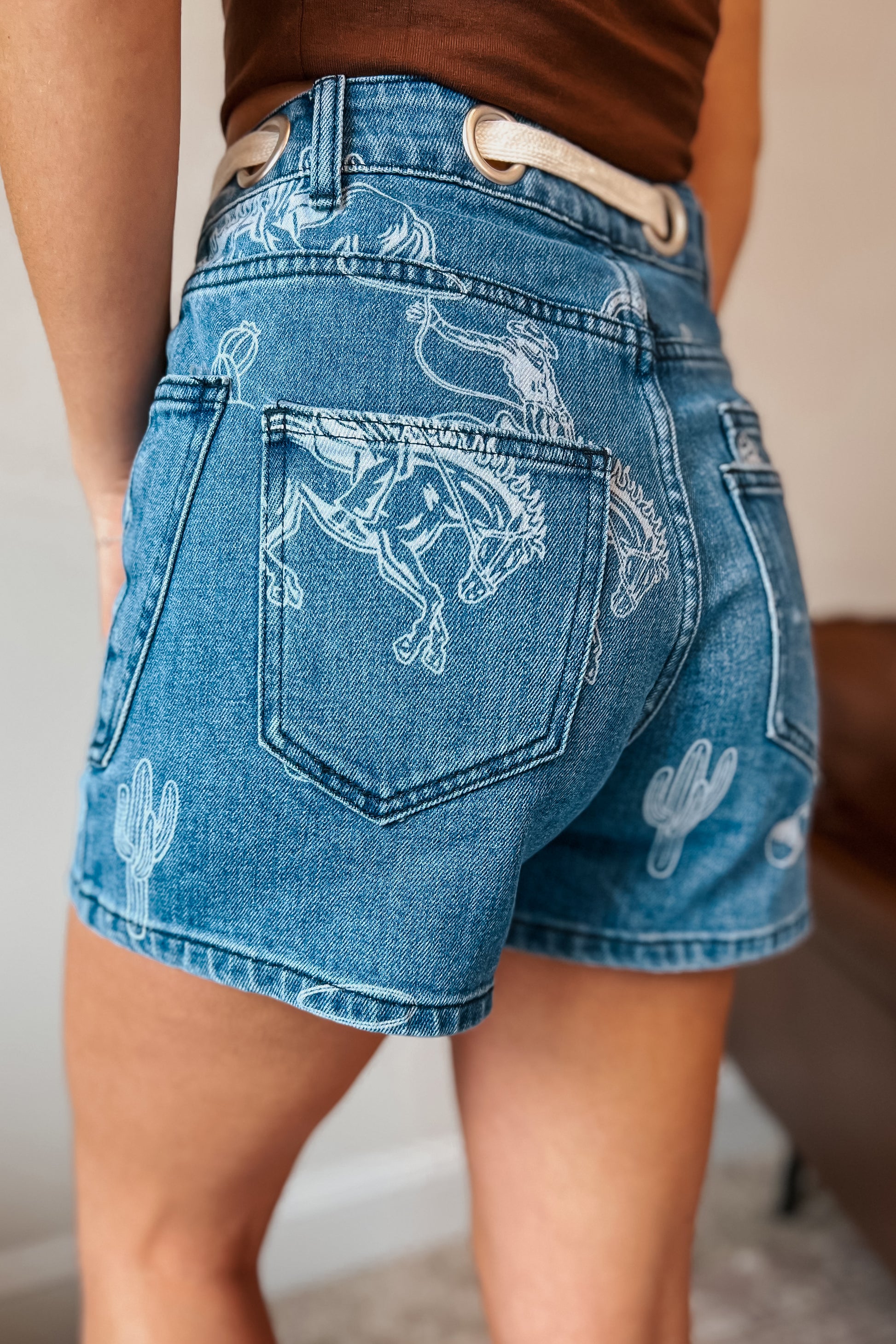 Medium wash denim shorts with bleached western print featuring bucking horses, cowboys, cacti, and horseshoes. High-rise fit with front and back patch pockets, belt loops, and cream rope drawstring threaded through large metal grommets at the waistband.