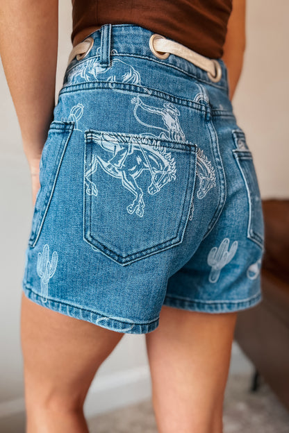 Medium wash denim shorts with bleached western print featuring bucking horses, cowboys, cacti, and horseshoes. High-rise fit with front and back patch pockets, belt loops, and cream rope drawstring threaded through large metal grommets at the waistband.