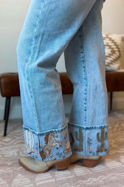 Close-up of a person wearing light blue wide-leg jeans with embroidered cowboy boots, showing the cuffed hems decorated with a horse and cactus pattern.