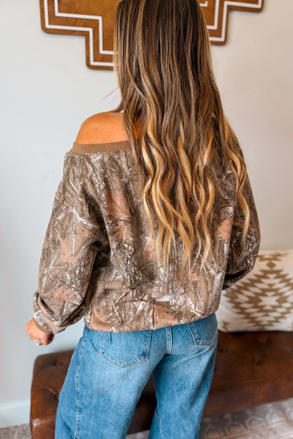 A woman viewed from behind wearing a brown camouflage off-the-shoulder long-sleeve crop top and blue jeans, standing indoors in front of a wall with a wooden mirror and a patterned cushion.