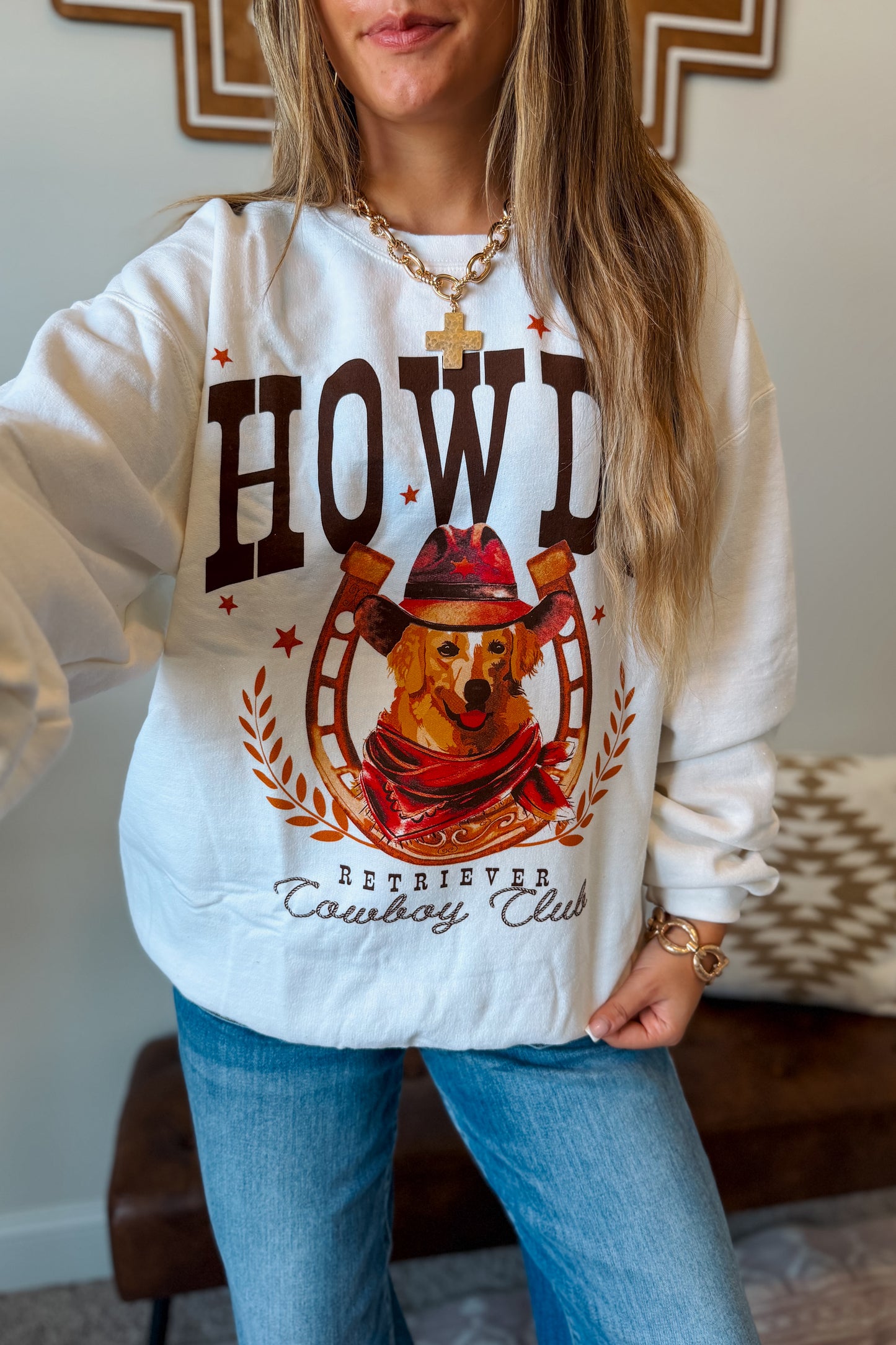 A woman wearing a white crewneck sweatshirt with a rustic cowboy dog graphic that reads 'HOWDY RETRIEVER Cowboy Club' with a dog in a hat inside a horseshoe, paired with blue jeans.
