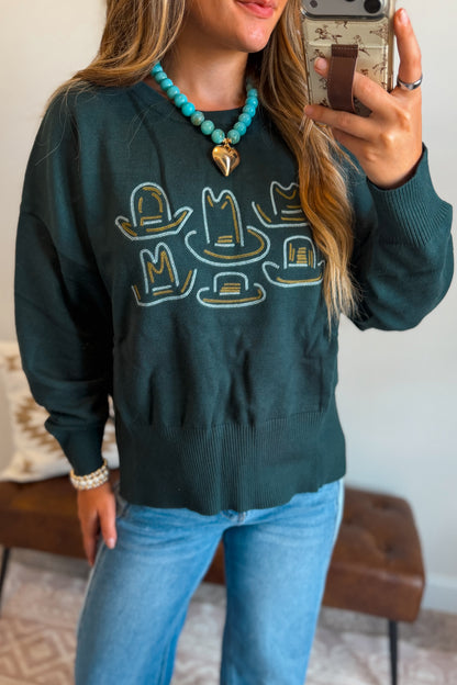 A woman takes a mirror selfie wearing a dark teal crewneck sweatshirt with embroidered cowboy hats, paired with light blue jeans and a turquoise beaded necklace.