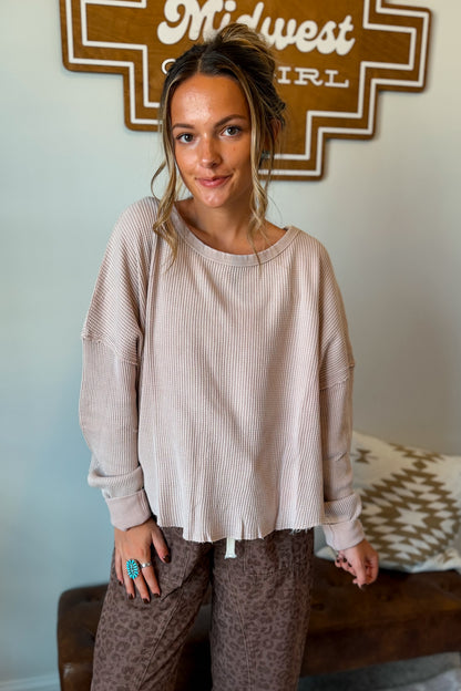 A woman wearing a beige ribbed long-sleeve top with raw, frayed hems and brown leopard-print pants, standing in front of a wall with a Midwest Girl sign.