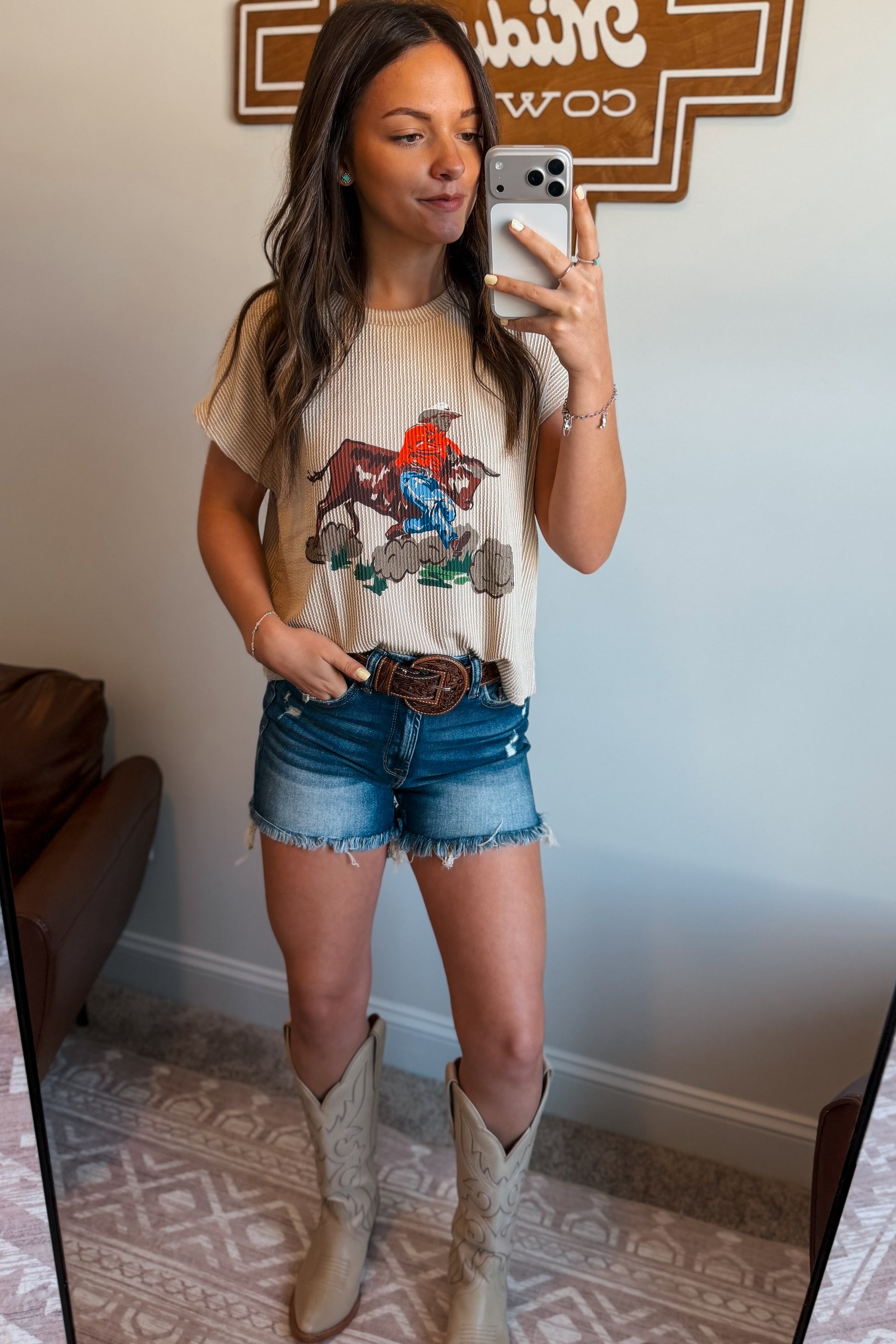 Cream ribbed short sleeve top featuring a western graphic of a cowboy riding a bucking bull, styled with denim shorts and a brown western belt