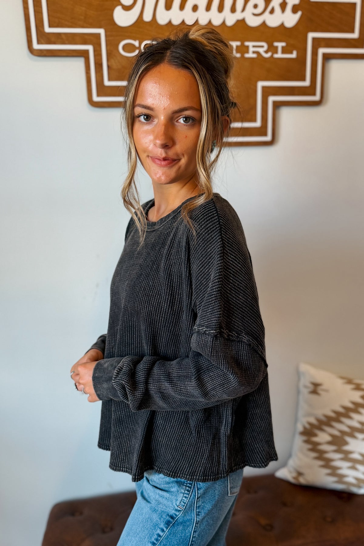 A woman wearing a dark charcoal gray ribbed long-sleeve top and light blue jeans, standing in a room with a wooden sign on the wall behind her.