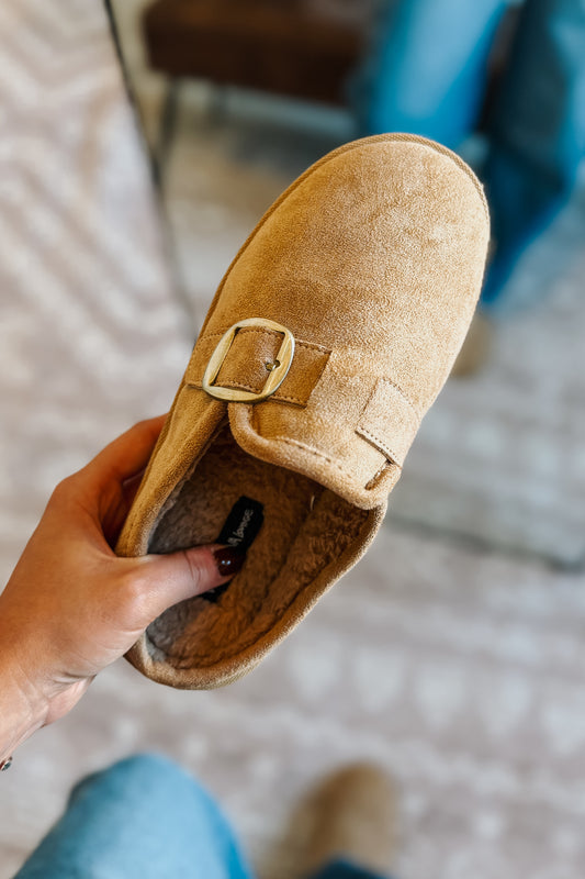 A tan suede mule slipper with a gold buckle strap, held in a hand against a blurred indoor background.