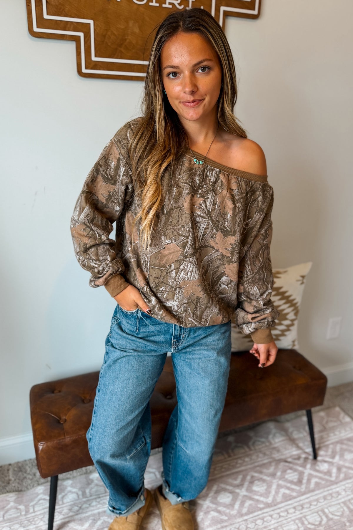 A woman wearing a brown and beige camouflage print off-the-shoulder long-sleeve top, blue jeans, and tan boots, standing in a room with a brown leather bench.