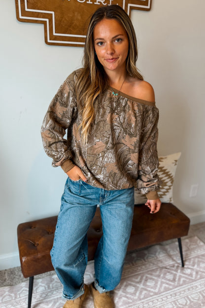 A woman wearing a brown and beige camouflage print off-the-shoulder long-sleeve top, blue jeans, and tan boots, standing in a room with a brown leather bench.