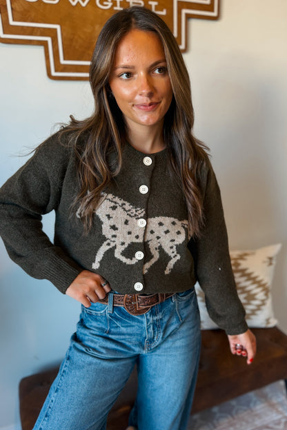 A woman wearing a dark olive green knit cardigan with a cream-colored dalmatian graphic on the front, paired with blue jeans and a brown belt, standing in an indoor setting.