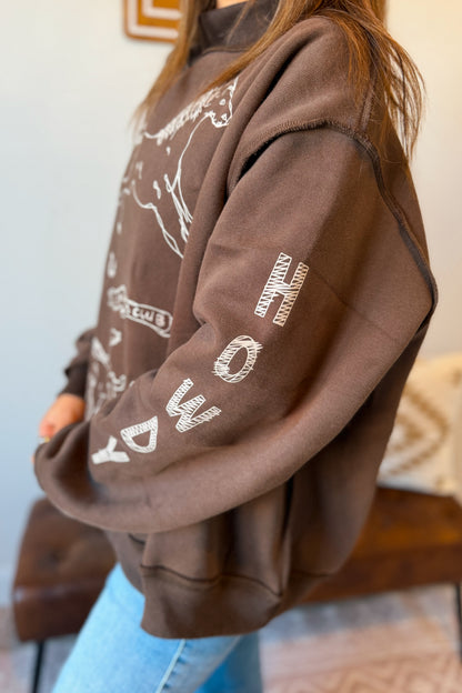 A person wearing an oversized brown sweatshirt with white graphic text and line art on the sleeves, paired with light blue jeans in an indoor setting.