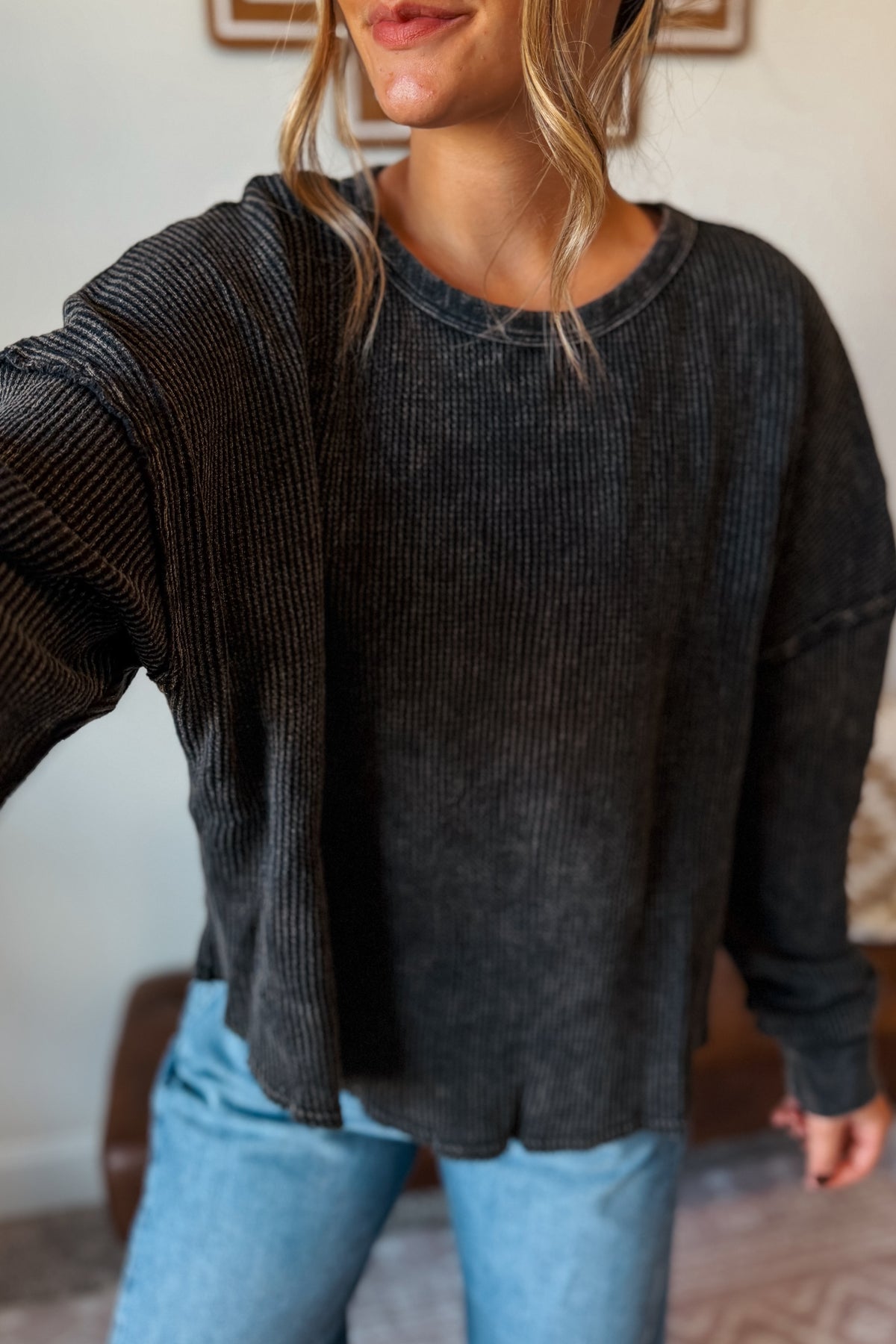 A woman wearing a dark charcoal gray ribbed knit crewneck sweater and light blue jeans, taking a mirror selfie in an indoor setting.