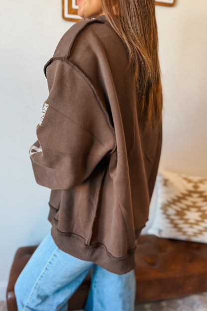 A woman wearing an oversized brown sweatshirt and light blue jeans, shown from a side-back angle in an indoor setting.
