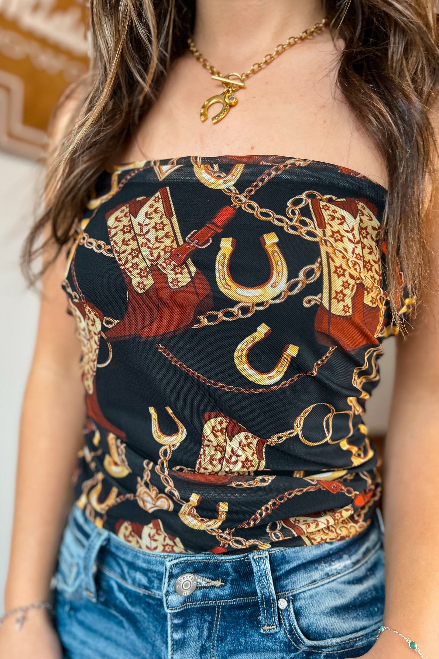 Strapless black western print top featuring cowboy boots, horseshoes, and gold chain details, shown styled with blue jeans.