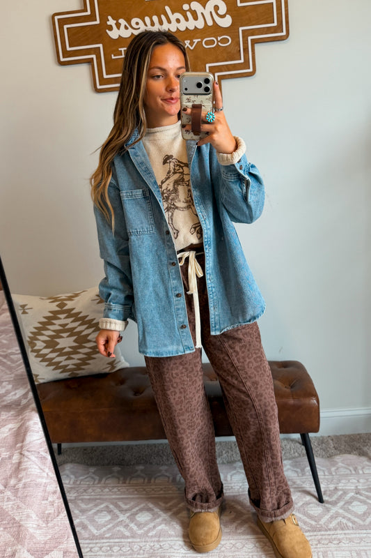A woman takes a mirror selfie showing an outfit consisting of a light blue denim jacket over a cream graphic t-shirt, paired with brown leopard-print pants and tan boots, in a bedroom setting.