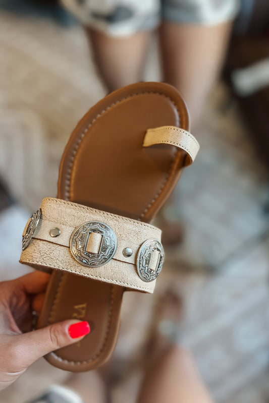 Cream western slide sandals with tan cushioned footbed and toe loop. Wide textured strap features silver engraved concho accents and metal studs. Brand name printed on footbed.