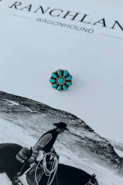 A small round turquoise flower-shaped stud earring with a gold-toned textured border, displayed on a white book 