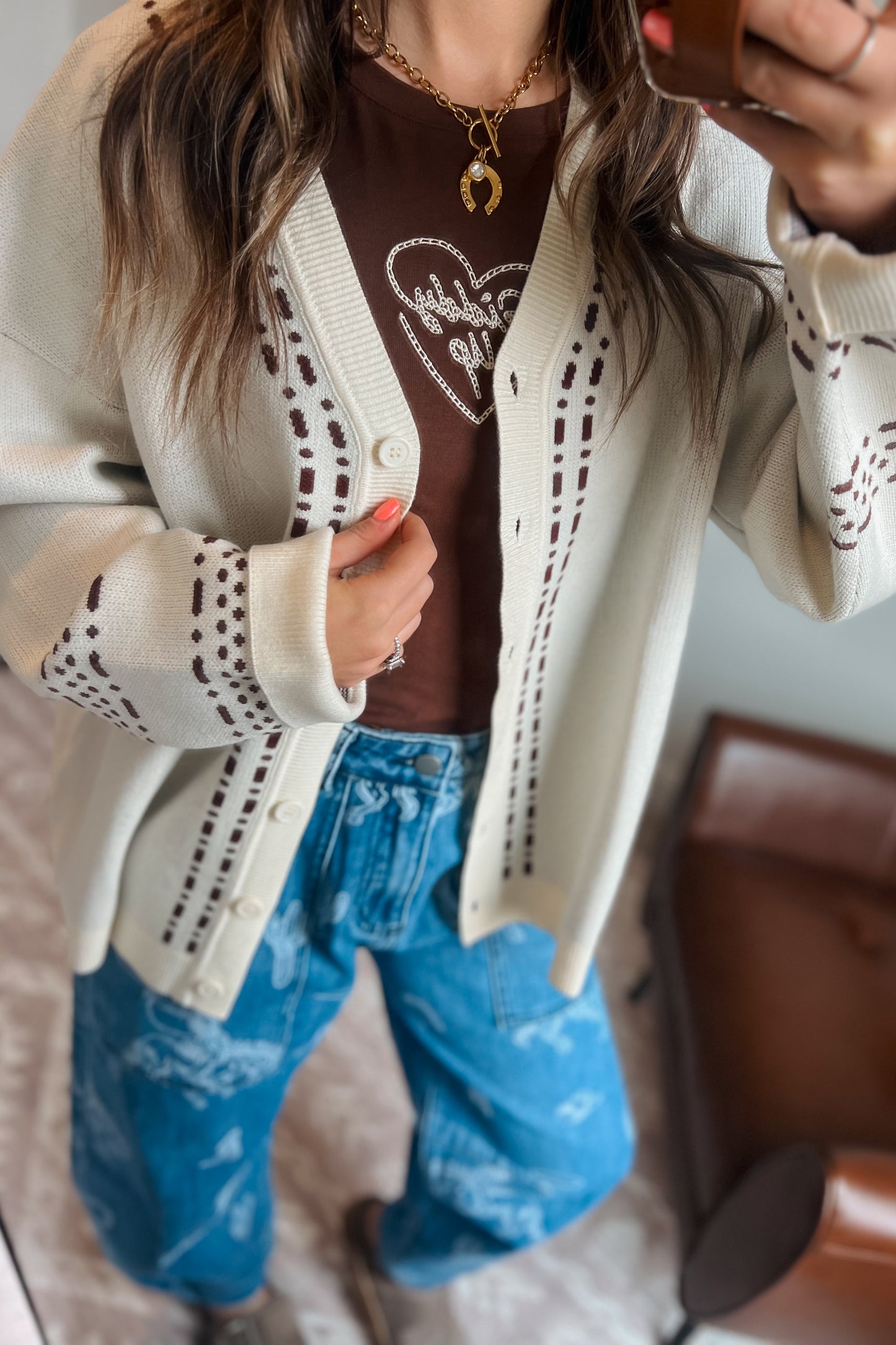 Model wearing a cream knit cardigan with brown western-inspired stitch detailing over a brown graphic tee and blue western print jeans.