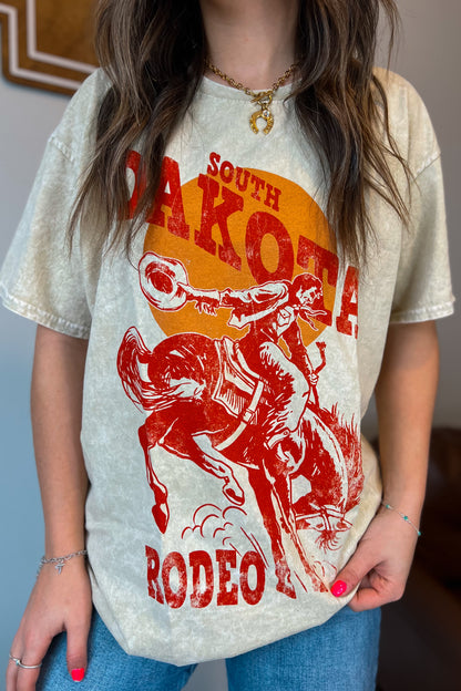 Light tan mineral-wash T-shirt with red “South Dakota Rodeo” graphic featuring a cowboy riding a bucking horse inside an orange sunset design