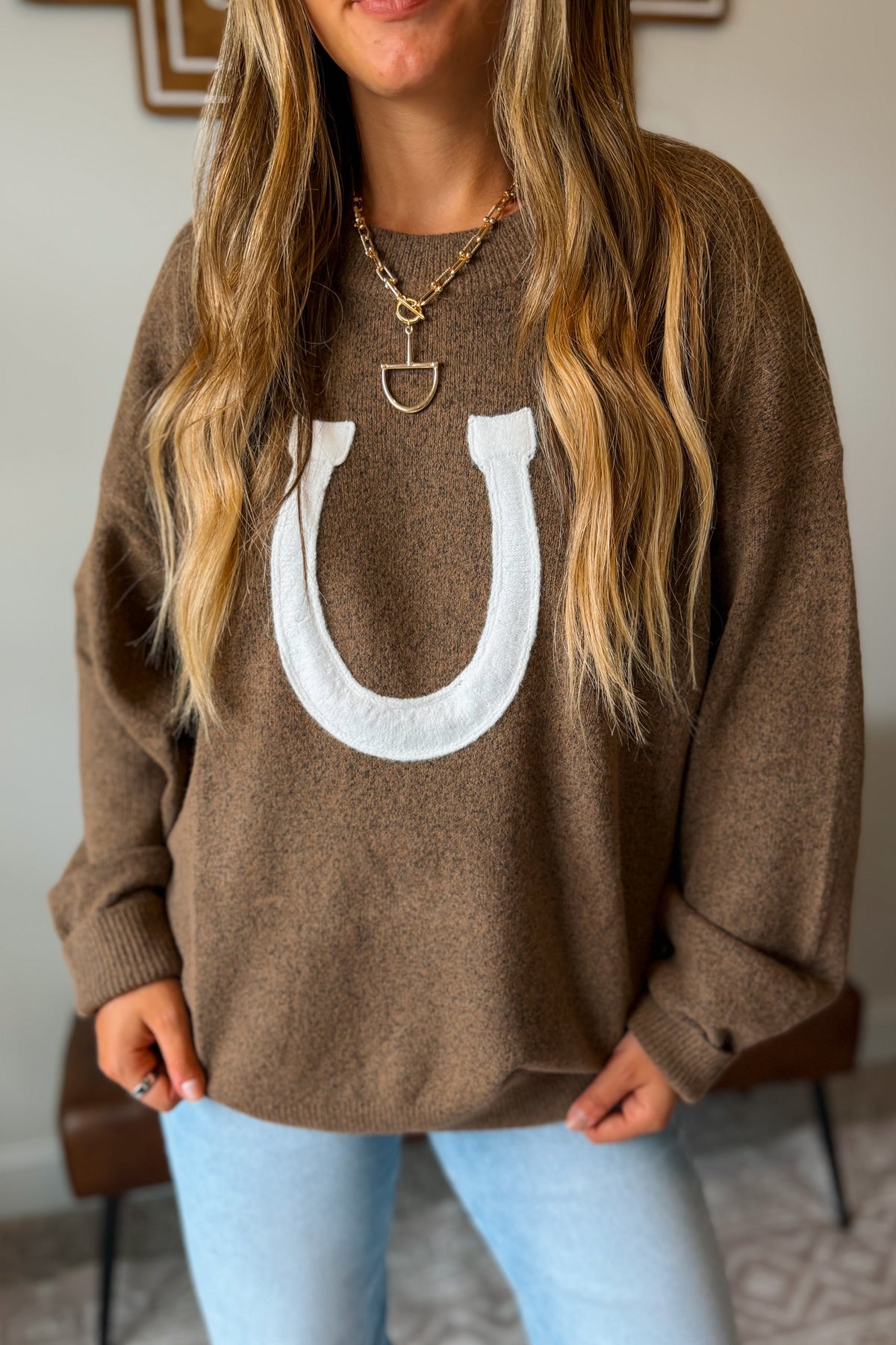 A woman wearing a brown knit crewneck sweater with a large white horseshoe graphic on the front, paired with light blue jeans and gold necklaces.