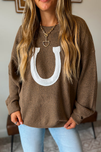 A woman wearing a brown knit crewneck sweater with a large white horseshoe graphic on the front, paired with light blue jeans and gold necklaces.