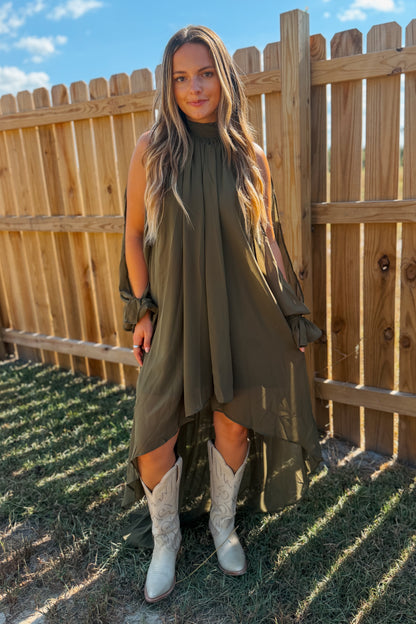 A woman stands outdoors wearing an olive green high-low dress with ruffled sleeves and light beige western boots.