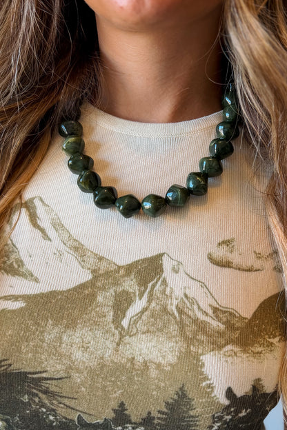 A close-up of a person wearing a dark green faceted bead necklace over a beige graphic t-shirt with a mountain print.