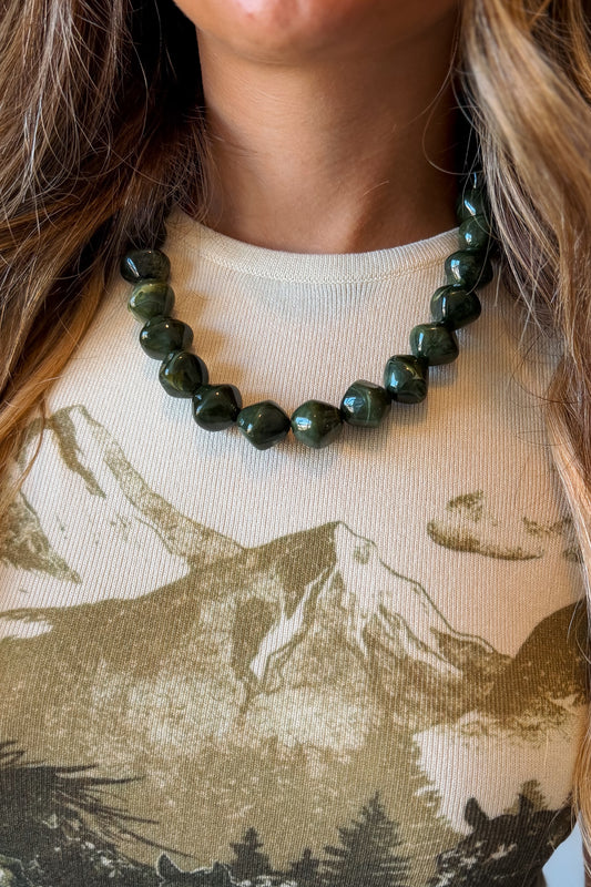 A close-up of a person wearing a dark green faceted bead necklace over a beige graphic t-shirt with a mountain print.