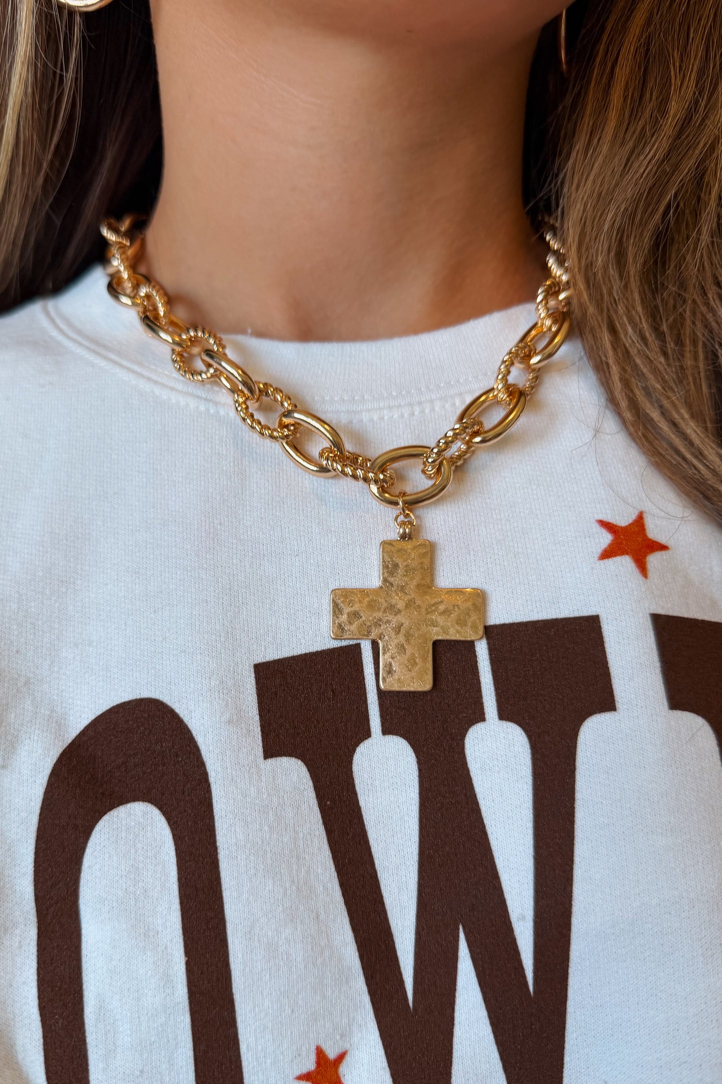 A close-up of a person wearing a chunky gold chain necklace with a textured gold cross pendant, paired with a white graphic t-shirt.