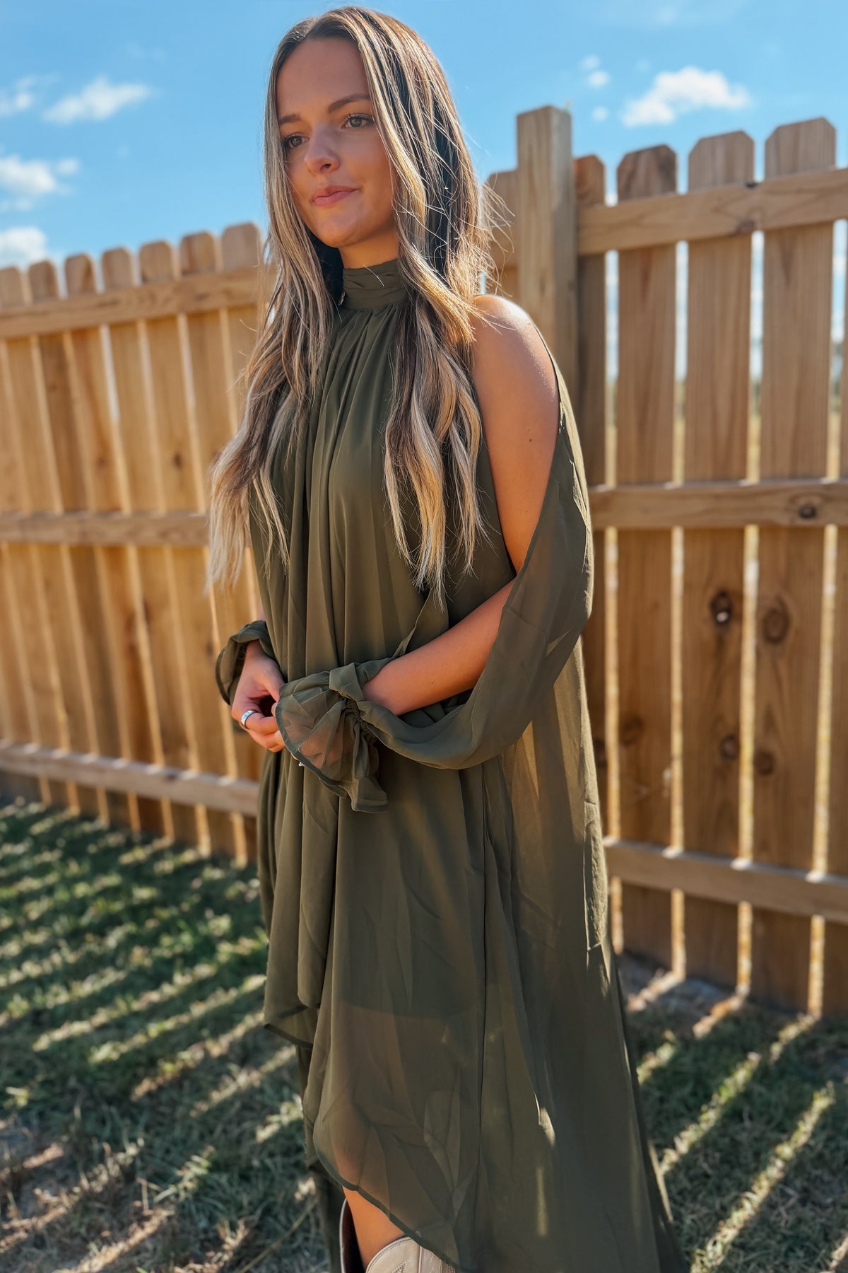 A woman wearing an olive green cold-shoulder midi dress with a high neck and tie detail at the waist, standing outdoors in front of a wooden fence under a blue sky.