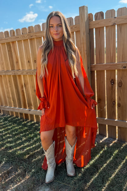 A woman wearing a rust-orange high-low dress with sheer sleeves and white cowboy boots, standing outdoors in front of a wooden fence.