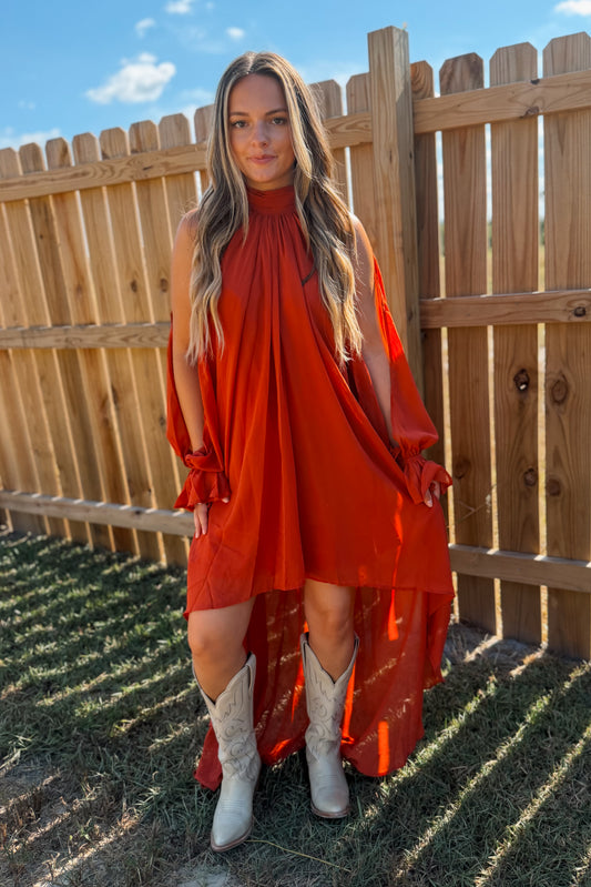 A woman wearing a rust-orange high-low dress with sheer sleeves and white cowboy boots, standing outdoors in front of a wooden fence.