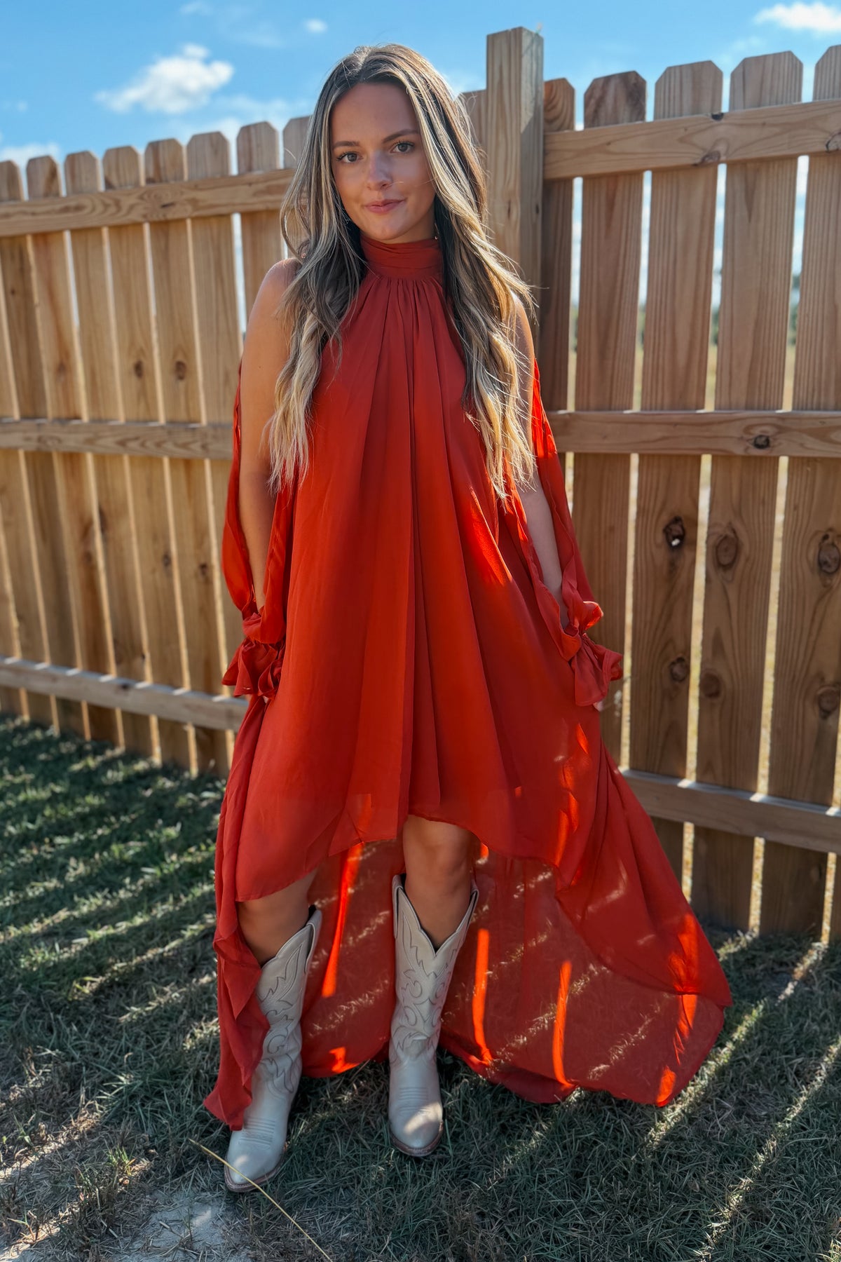 A woman stands outdoors in front of a wooden fence wearing a flowing rust-orange high-low dress with long sleeves and white cowboy boots.