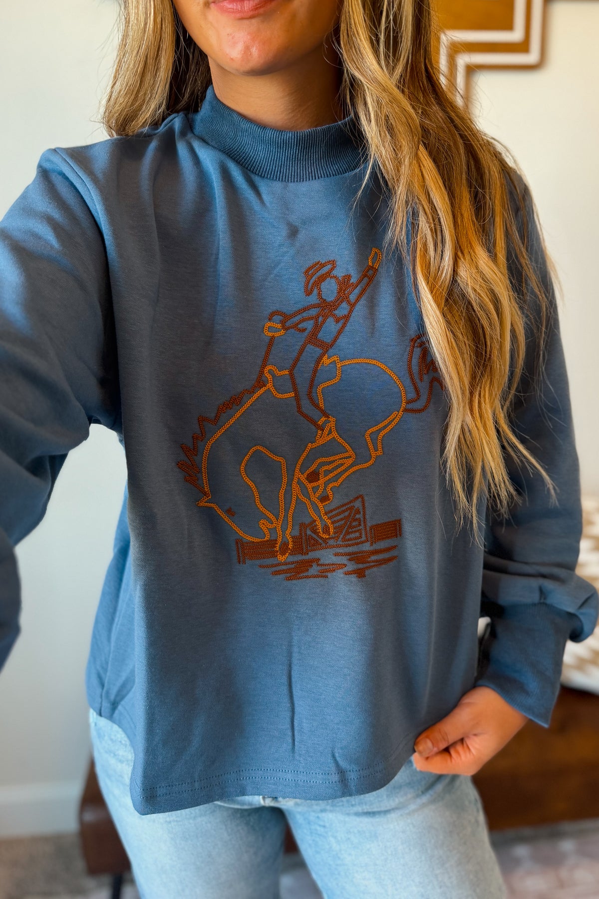 A woman wearing a dusty blue long-sleeve mock neck sweatshirt with a brown graphic of a cowboy riding a bucking bull, paired with light wash jeans.