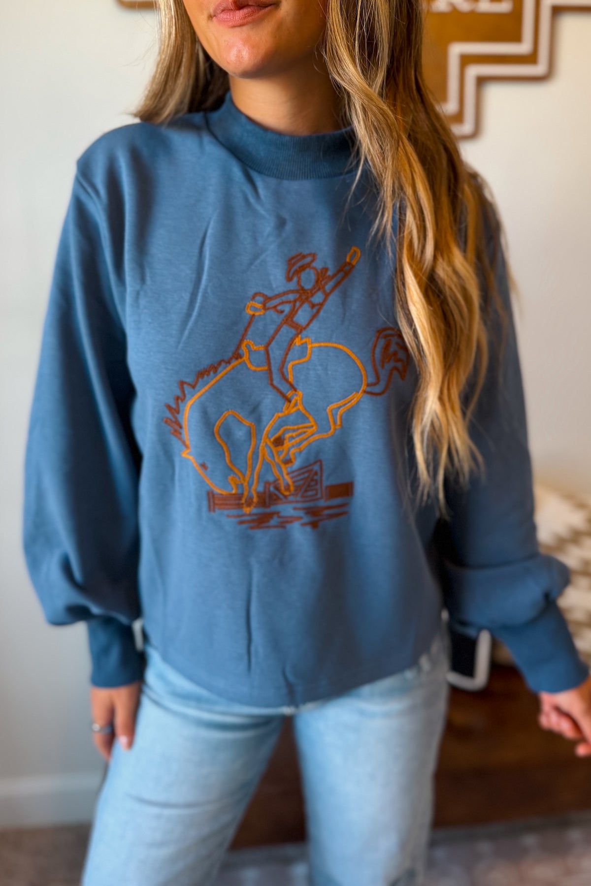 A woman wearing a dusty blue sweatshirt with an orange graphic of a cowboy riding a bucking horse, paired with light-wash jeans.