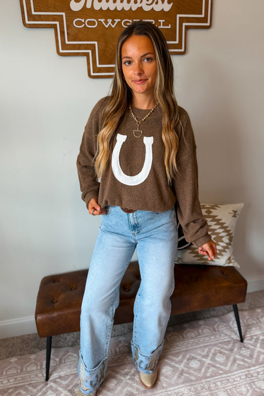 A woman wearing a brown sweater with a large white horseshoe graphic, paired with light-wash blue jeans and western boots.