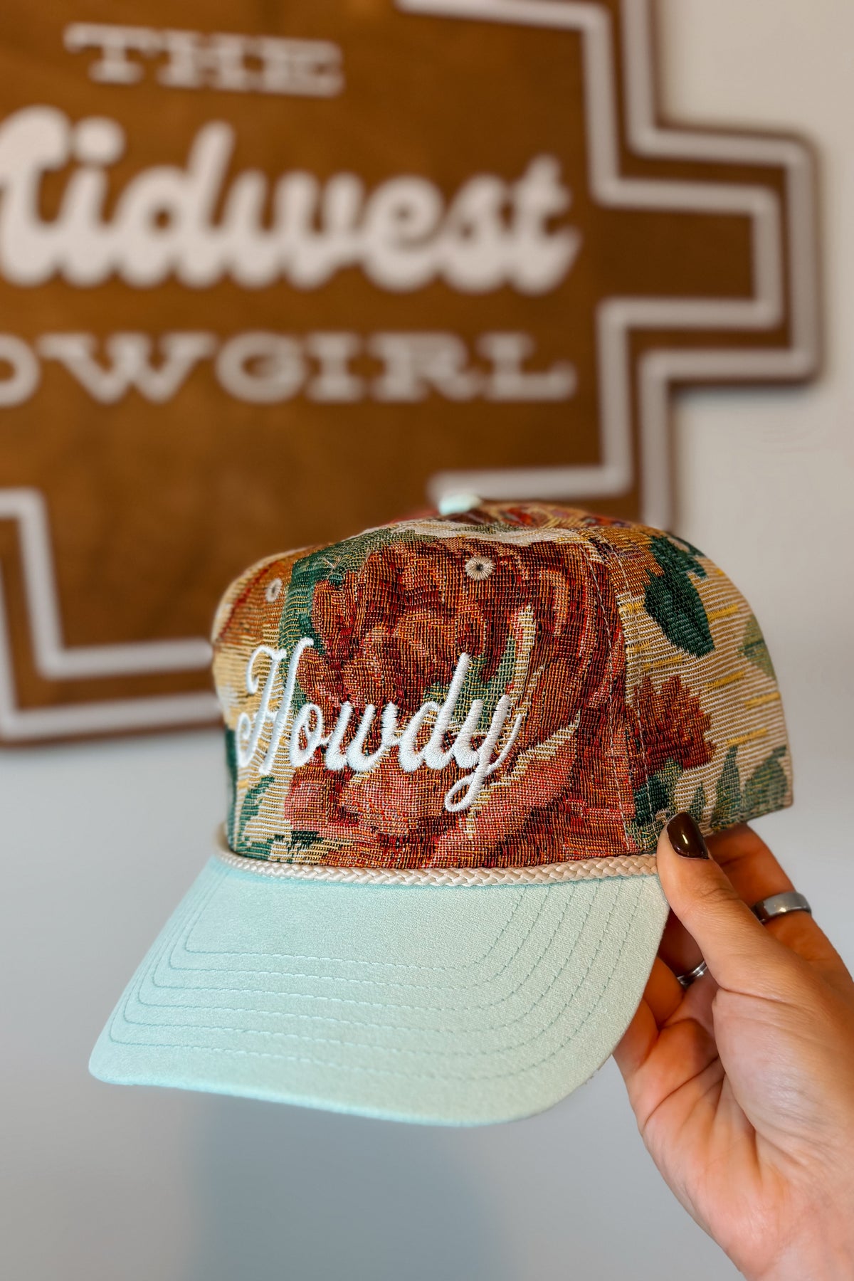 A hand holds a baseball cap with a floral front panel, white cursive 'Howdy' embroidery, and a light mint green brim. A brown sign reading 'The Midwest Cowgirl' is blurred in the background.