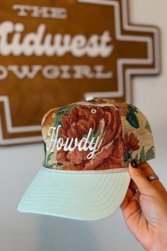 A hand holds a baseball cap with a floral front panel, white cursive 'Howdy' embroidery, and a light mint green brim. A brown sign reading 'The Midwest Cowgirl' is blurred in the background.