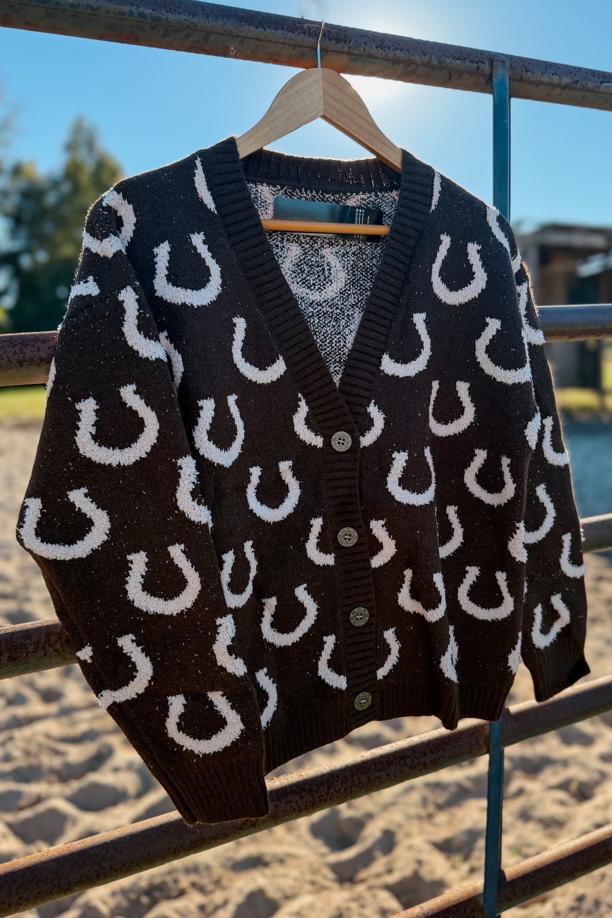 A dark brown knit cardigan with a white horseshoe pattern, hanging on a wooden hanger from an outdoor metal railing.