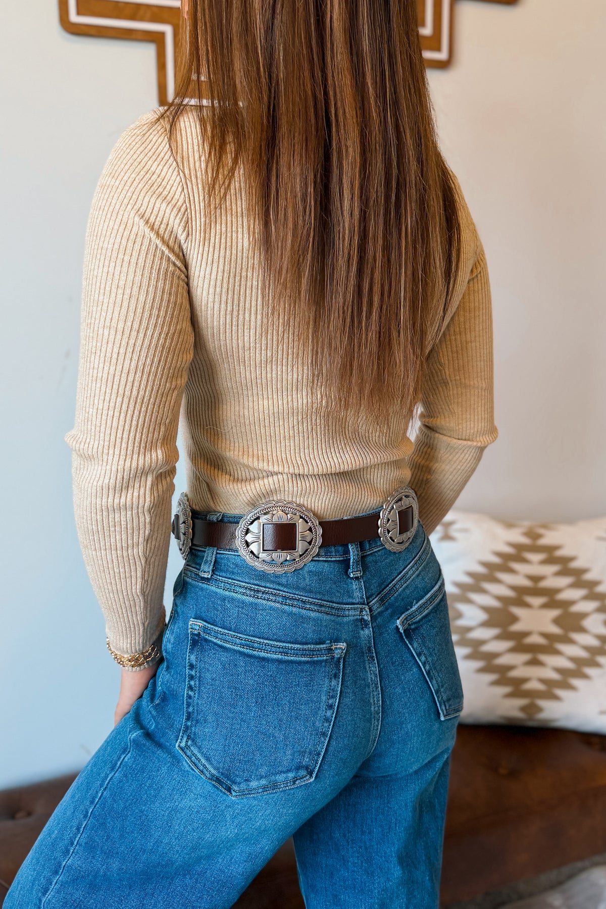A woman shown from the back wearing a beige ribbed long-sleeve top, blue jeans, and a brown leather belt with large silver concho buckles, in an indoor setting.
