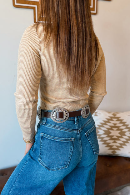 A woman shown from the back wearing a beige ribbed long-sleeve top, blue jeans, and a brown leather belt with large silver concho buckles, in an indoor setting.