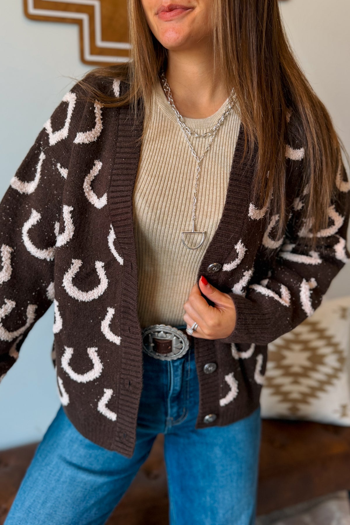 A woman wearing a dark brown open-front cardigan with a white horseshoe pattern, layered over a beige ribbed sweater and blue jeans.