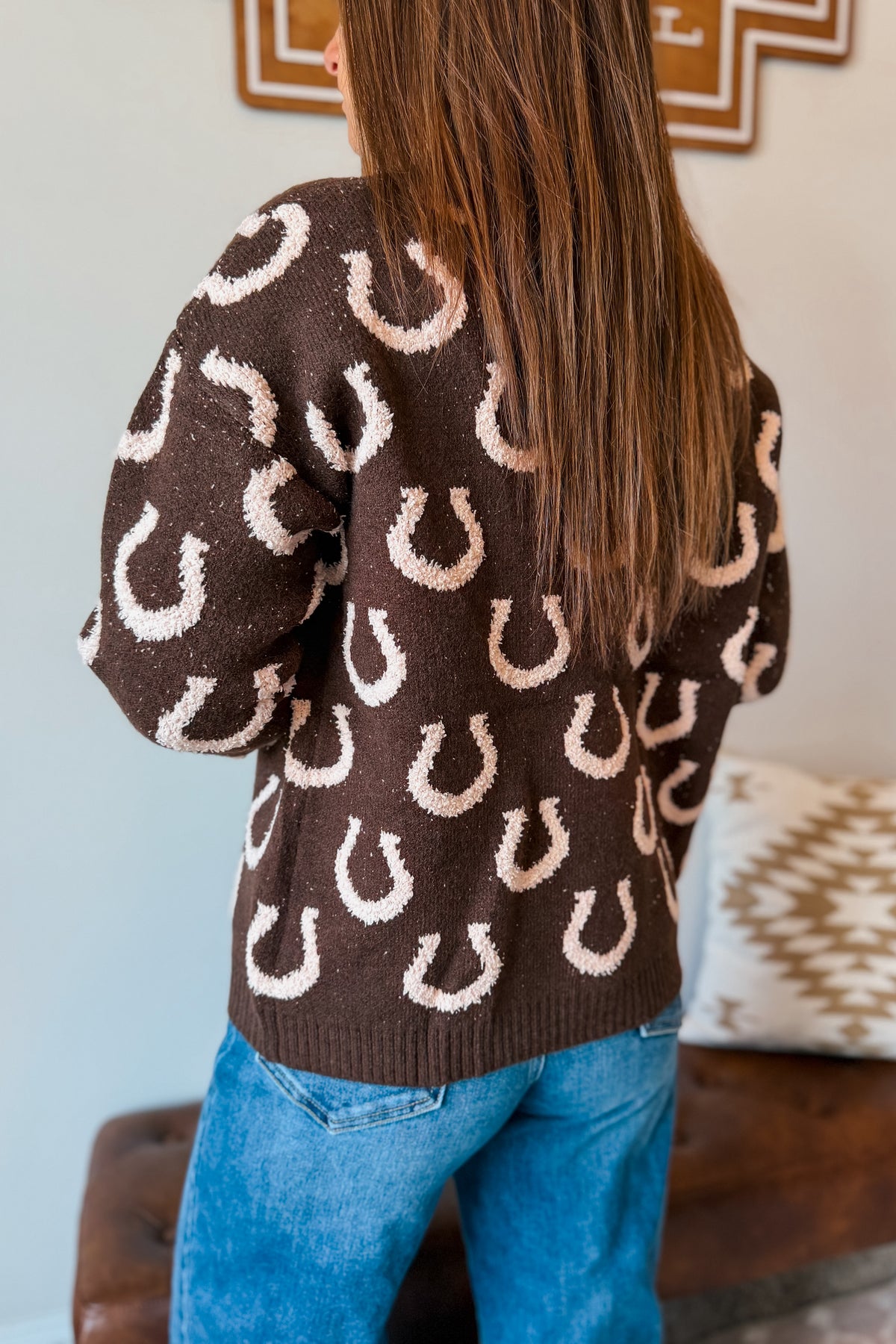 A person shown from the back wearing a brown knit sweater with a repeating white horseshoe pattern, paired with blue jeans in an indoor setting.