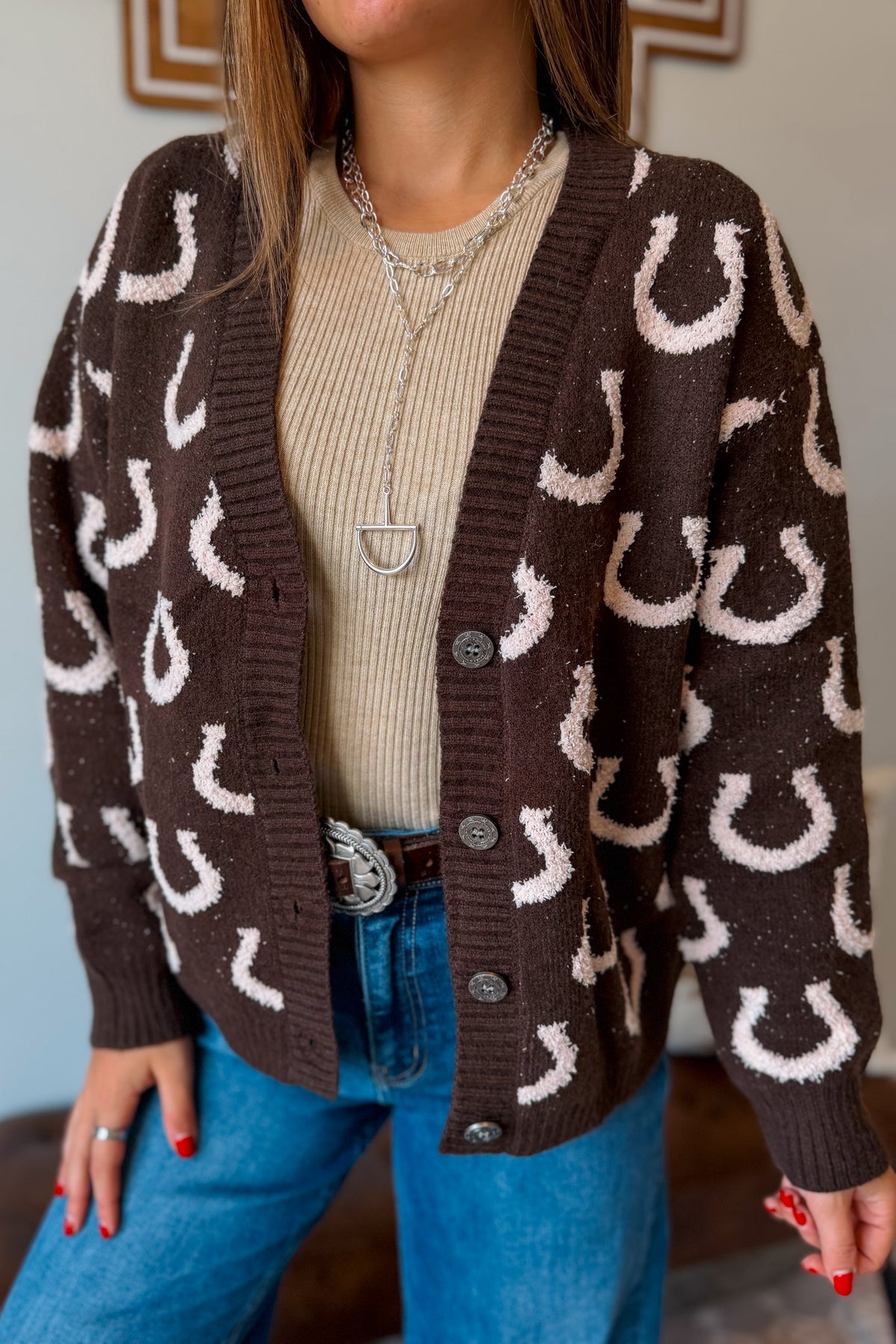 A woman wearing a brown knit cardigan with a white horseshoe pattern, layered over a beige ribbed sweater and blue jeans.