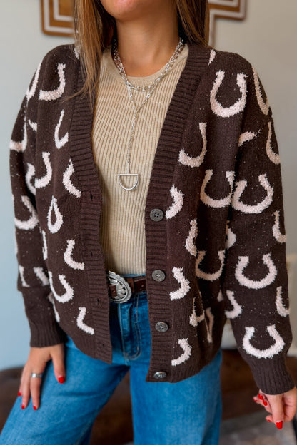 A woman wearing a brown knit cardigan with a white horseshoe pattern, layered over a beige ribbed sweater and blue jeans.